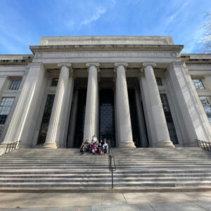 Group of students sitting on the front steps of the Massachusetts Institute of Technology (MIT) under the tall columns of the iconic main entrance. They were part of the Boston Bootcamp.
