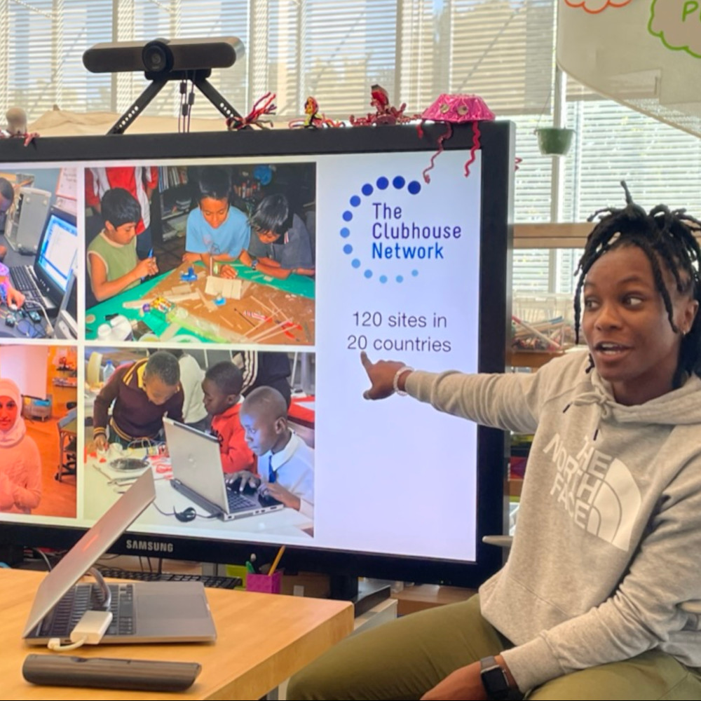 A person pointing to a presentation slide that reads "The Clubhouse Network: 120 sites in 20 countries," with images of diverse children working on technology and creative projects.